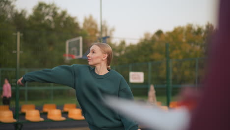 close-up blurred view of someone observing lady in green hoodie serving volleyball on outdoor court, with background featuring people near football field and orange stadium seats