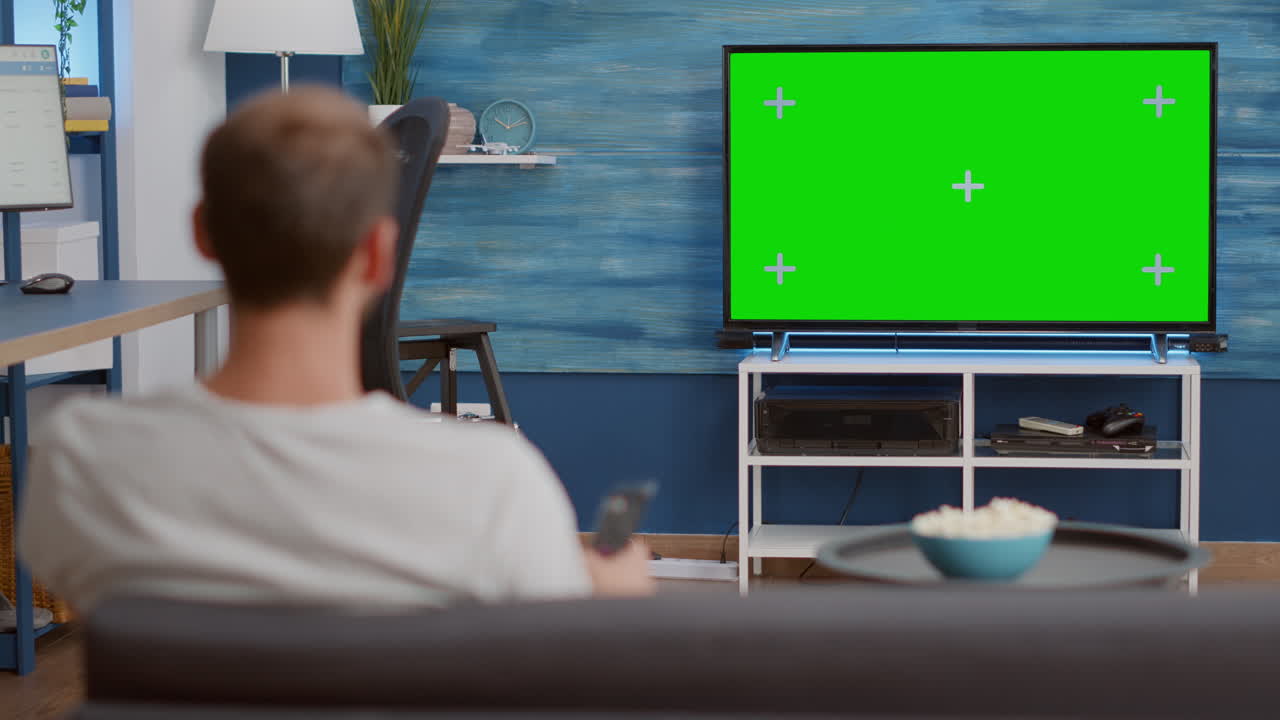 Static tripod shot of man switching channels while looking at green screen on tv