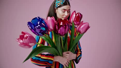 fashion model wearing colorful striped suit and headband holding bouquet of colorful tulips posing on pink background for spring or summer campaign