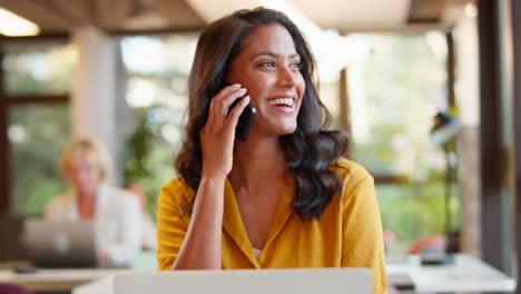 Mature-Businesswoman-Working-On-Laptop-At-Desk-In-Office-Taking-Call-On-Mobile-Phone