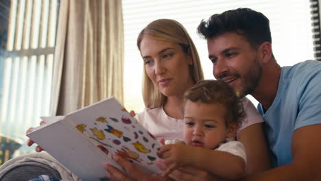 family sitting on sofa at home with parents reading book with young son