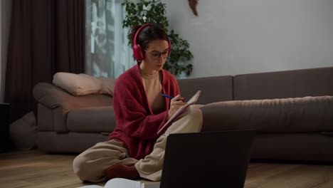 a confident and serious brunette girl with glasses in wireless red headphones and a sweater sits on the floor in front of a gray laptop and writes down her notebooks for a foreign language lesson online