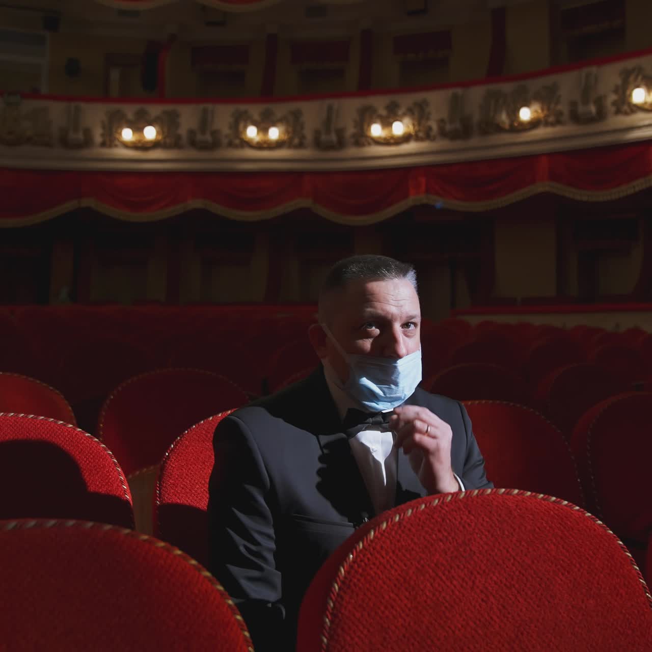 Alone businessman in mask in theater. Man in protective face mask sitting in a chair of empty auditorium. Theater in quarantine. Coronavirus pandemic.