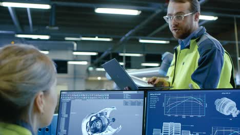 at the factory: male and female mechanical engineers use laptop and personal computer while discussing robotic arm manufacturing project. screen shows 3d engine concept made with cad software.