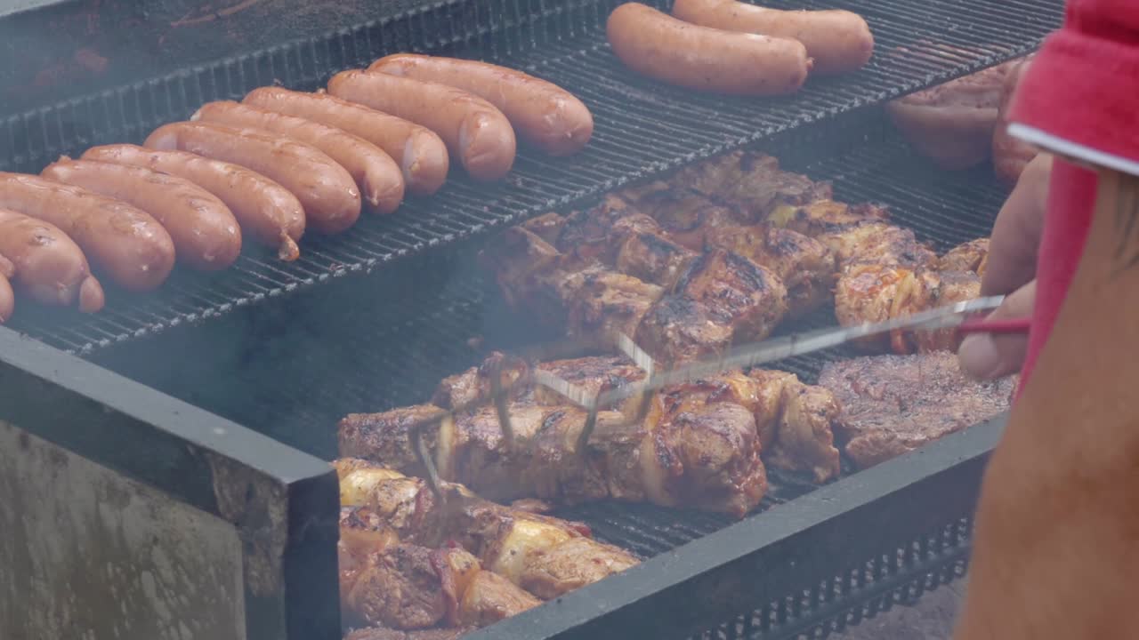 Man in the red T-shirt turns over the meat on the grill. He work outside.