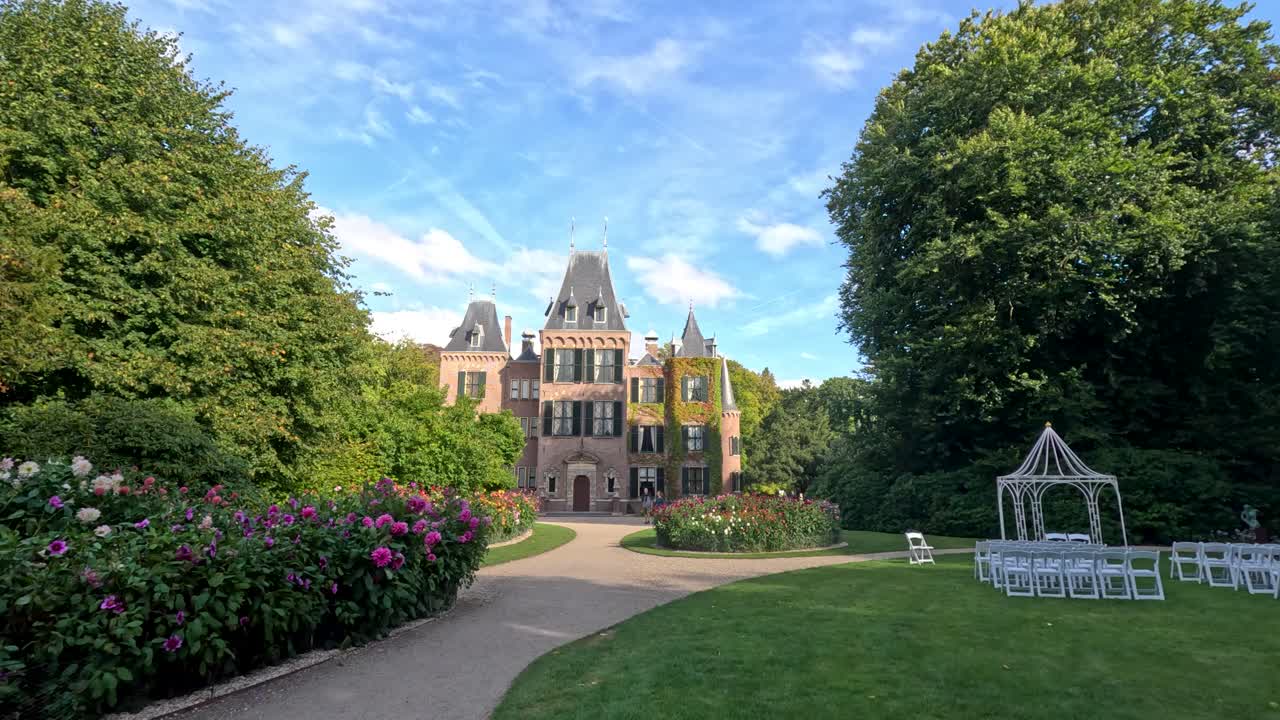 Wide-angle footage pans along a garden pathway toward a historic castle, surrounded by lush greenery, blooming flowers, and outdoor seating under bright daylight