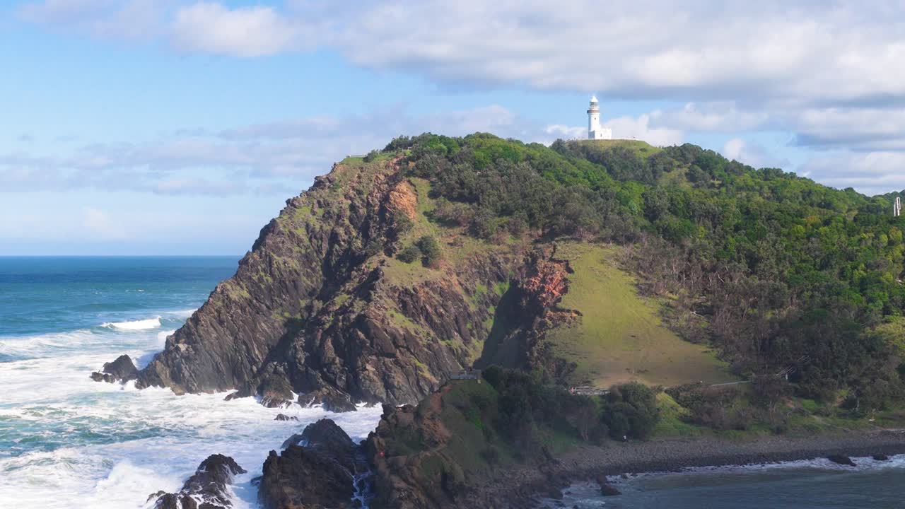 Aerial footage captures Byron Bay's lighthouse atop a lush cliff, surrounded by ocean waves under clear skies