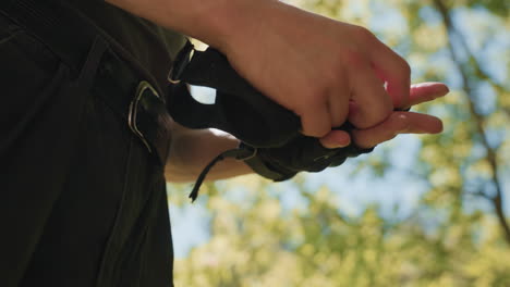 blurry view of green leafy canopy and clear blue sky framing young man in pants pulling off black fingerless tactical gloves at waist, hands grasping glove strap, sunlight filters through branches