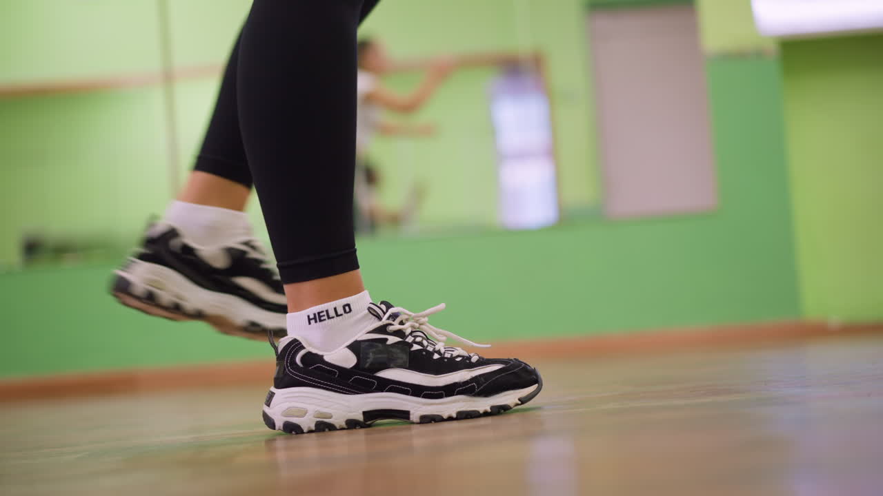 Leg view of woman in black leggings and canvas sneakers bouncing on polished wooden floor with mirror reflection showing blurred figure, capturing motion, rhythm, and dynamic indoor workout energy