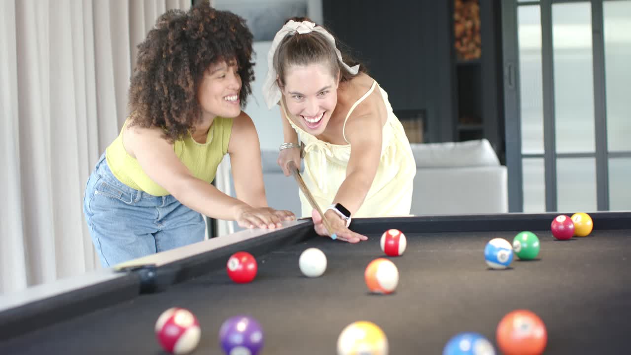 Amiche di diverse etnie che guidano la stecca e colpiscono la bilia bianca a un tavolo da biliardo a casa, festeggiando un gran tiro.