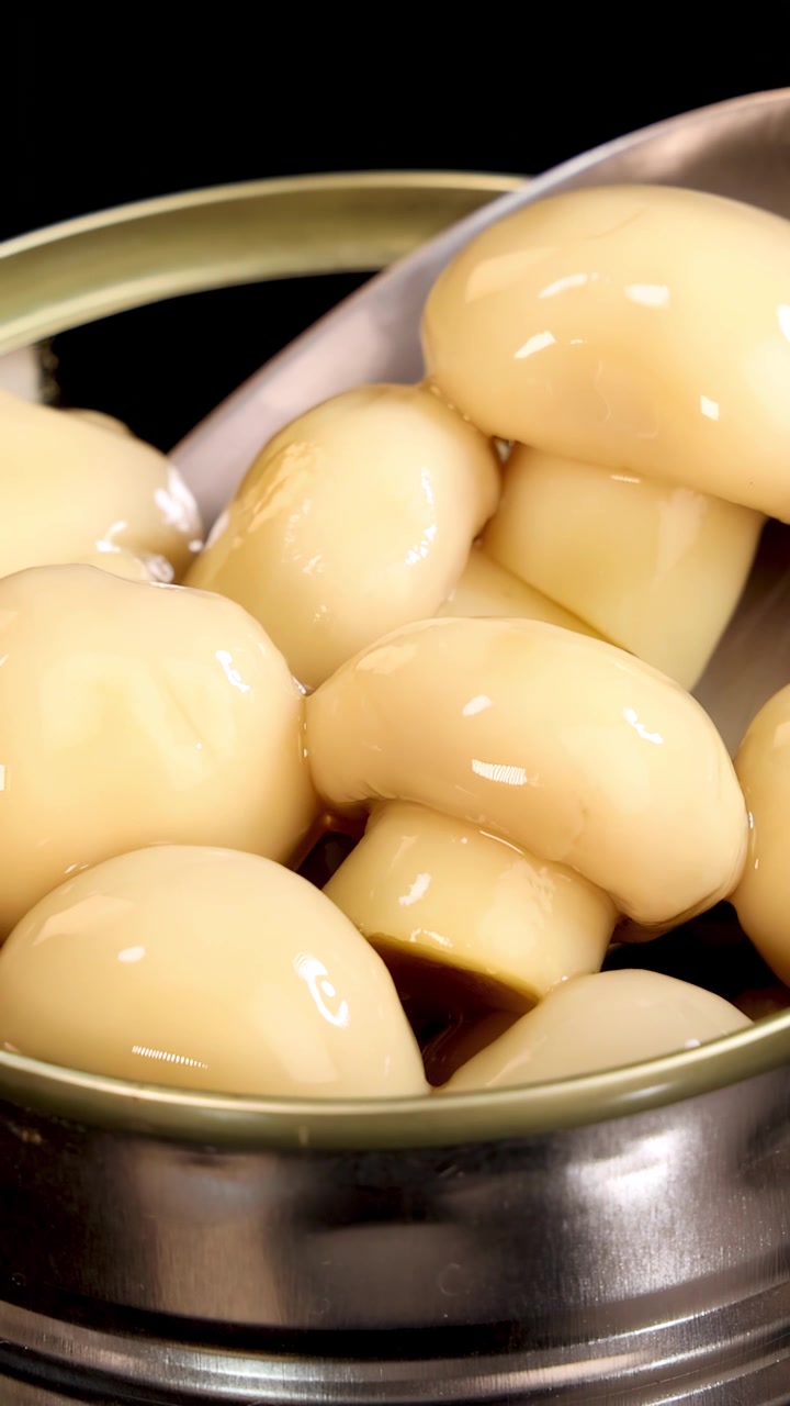 Una lattina di funghi champignon interi viene aperta e prelevata con un cucchiaio sotto una brillante illuminazione da studio, evidenziando la texture lucida e l'aspetto conservato