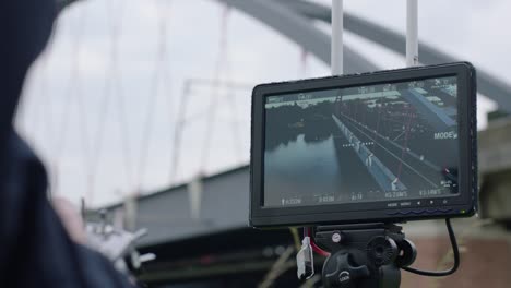 view from handheld camera of operator focusing tripod-mounted display on blue-arch bridge with red cables, with settings visible on connected monitor