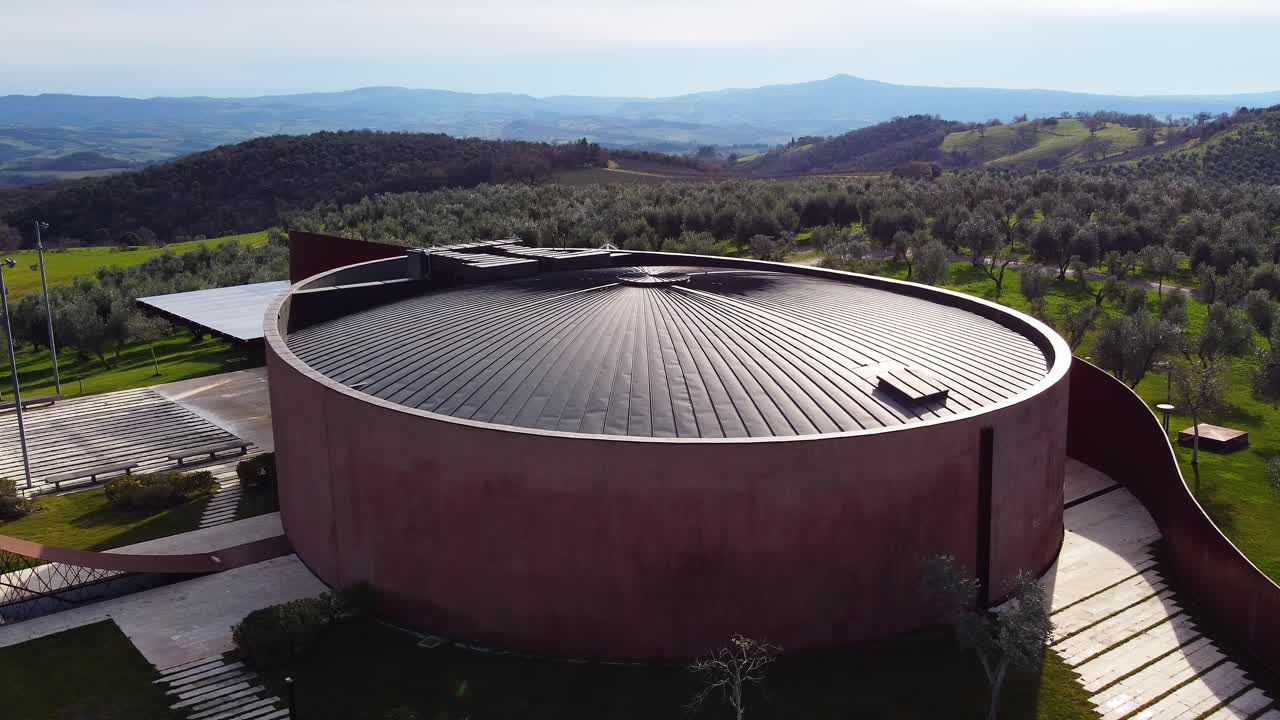 This aerial footage of Montecucco captures the Forum Fondazione Bertarelli surrounded by trees and vineyards. The camera performs a dolly movement to the right, revealing the scenic surroundings.