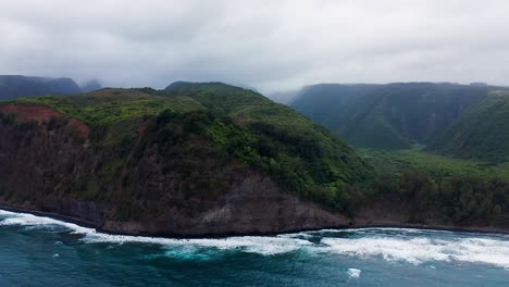 steep cliffs draped in lush green vegetation curve along the coastline as foamy blue waves churn below, while heavy gray clouds drift over this rugged and remote natural landscape.