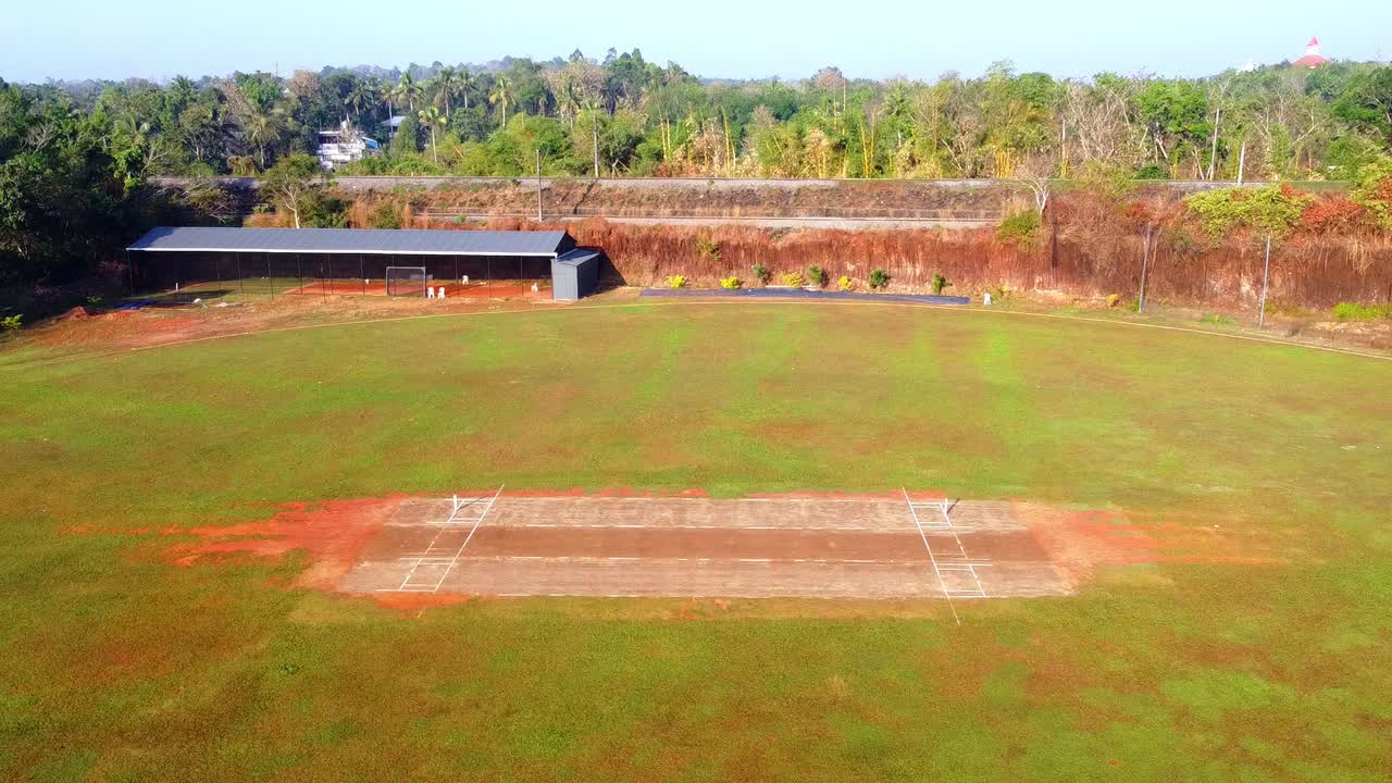 Inquadratura aerea con drone di un campo da cricket rurale con un campo da gioco in primo piano e un treno passeggeri che passa su un binario sopraelevato sullo sfondo.