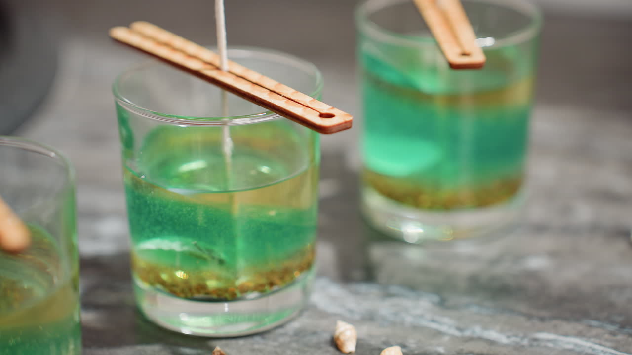 Three glass jars filled with layered green and yellow beeswax mixture, each containing upright wick secured by wooden wick stickers on top, while female hand drops shell-like decoration on the table