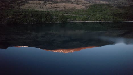 flying-backwards-to-reveal-the-perfectly-reflected-snow-capped-mountains-at-sunrise-in-Lake-Wakatipu-in-Queenstown-New-Zealand