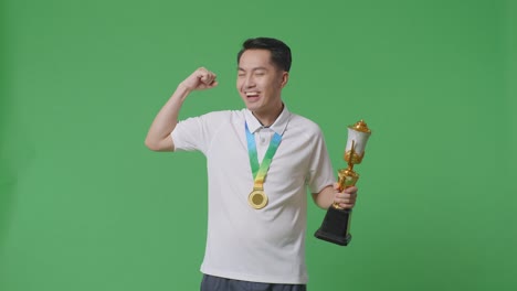 asian man with a gold medal and trophy flexing his bicep and smiling to camera on green screen background in the studio