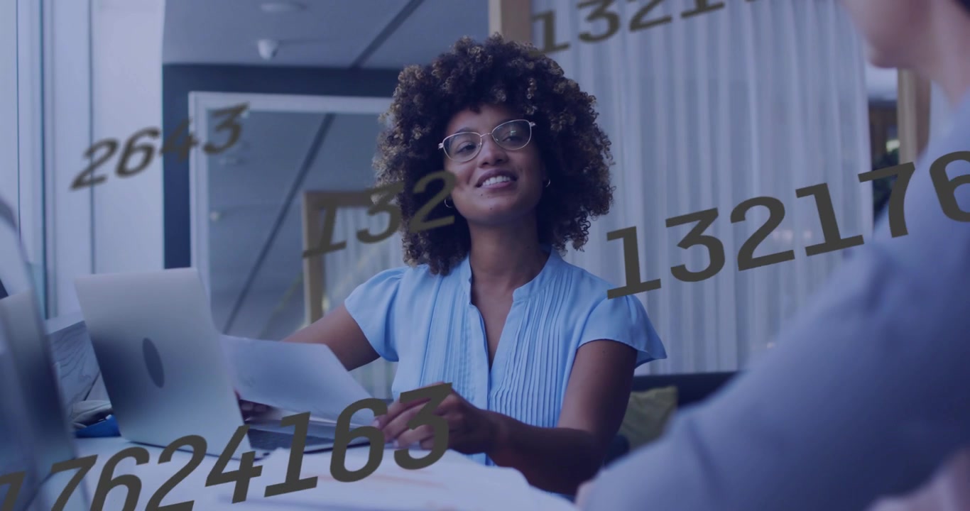 Mulher lendo documentos, levantando uma folha e verificando o laptop, explicando finanças enquanto números em movimento marcam as páginas.