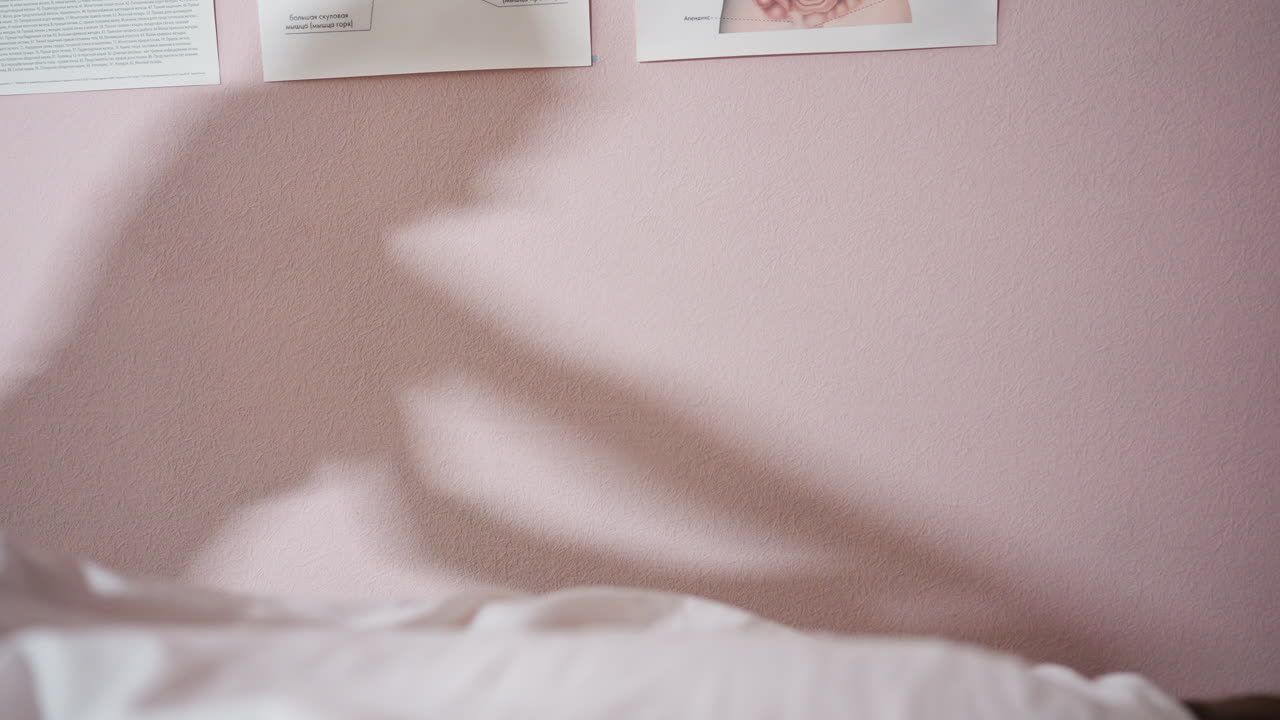 Shadow of massage therapist and client's interaction softly cast on pink wall during massage session, with slight visibility of anatomical charts above and white sheet covering client in foreground
