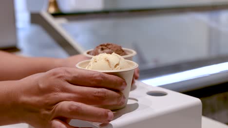 a hand lifts a paper cup of ice cream from a white display stand in a brightly lit dessert shop, with smooth camera movement and clear focus