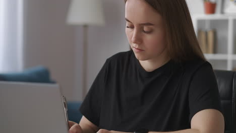 female-student-is-doing-homework-writing-notes-in-notebook-and-viewing-webinar-on-display-of-laptop-e-learning-and-distant-education