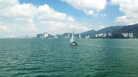 Velero-Navegando-En-El-Océano-Turquesa-En-Malasia-Camino-A-Langkawi-Con-Rascacielos-De-La-Ciudad-Y-Edificios-Altos-En-El-Fondo