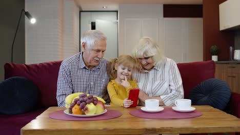 Grandparents-teaching-small-kid-granddaughter-using-smartphone-making-online-shopping-at-home