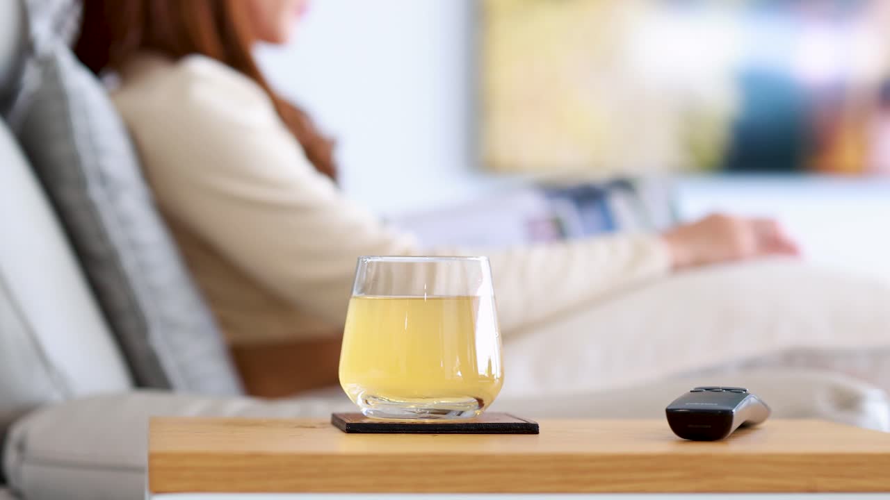An Asian woman sits on a sofa, reading a book and occasionally watching TV. A glass of tea and remote rest on a wooden tray. Bright, soft lighting creates a calm, modern living room atmosphere