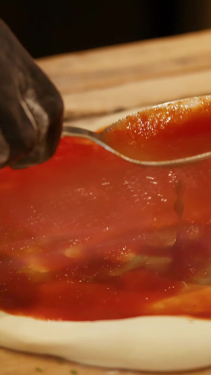 Primo piano di mani guantate che spalmano salsa di pomodoro rossa su impasto crudo per pizza in cucina.