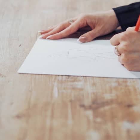 Dressmaker is drawing a creative design of a modern clothing indoors. Female's hands of a young tailor on the wooden table with a pencil. Feminine hobby.