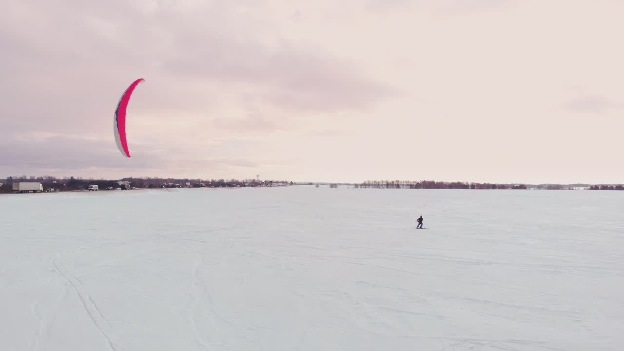 carrera de competencia de kite de nieve de deportes extremos de invierno aéreo de 4k con diferentes cometas de nieve coloridas, esquí, practicantes de snowboard sobre el lago de hielo frente a la ciudad en ventisca y clima de nieve desde drones