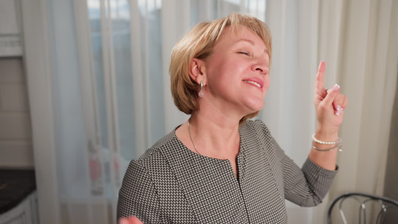 Cheerful housewife with short blond hair and painted lips dancing joyfully indoors in bright cozy home environment with natural daylight streaming through white curtains