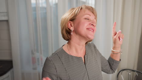 cheerful housewife with short blond hair and painted lips dancing joyfully indoors in bright cozy home environment with natural daylight streaming through white curtains