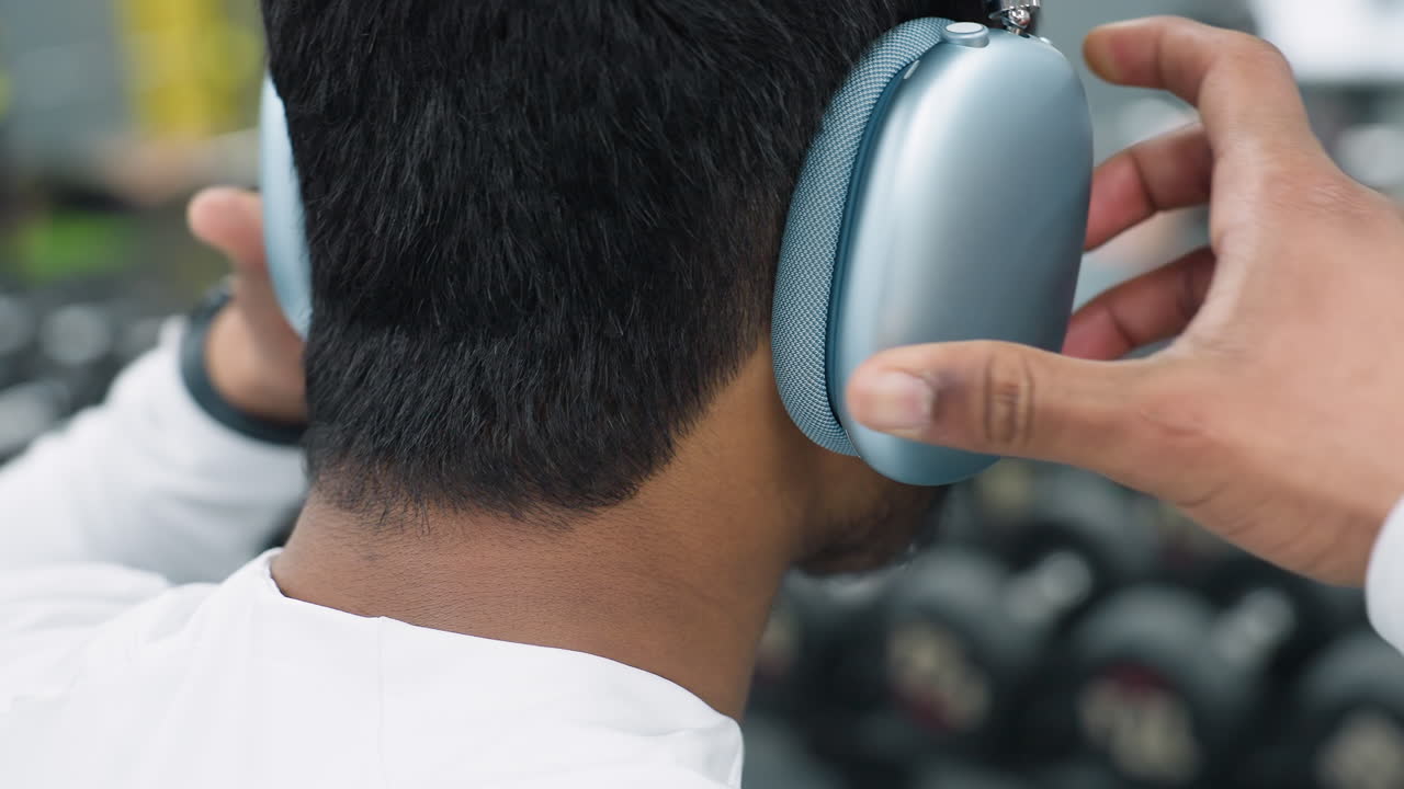 close up of young man putting on headset while turning neck in gym environment with blurred equipment and people in background, showcasing focused motion and tech use during workout preparation