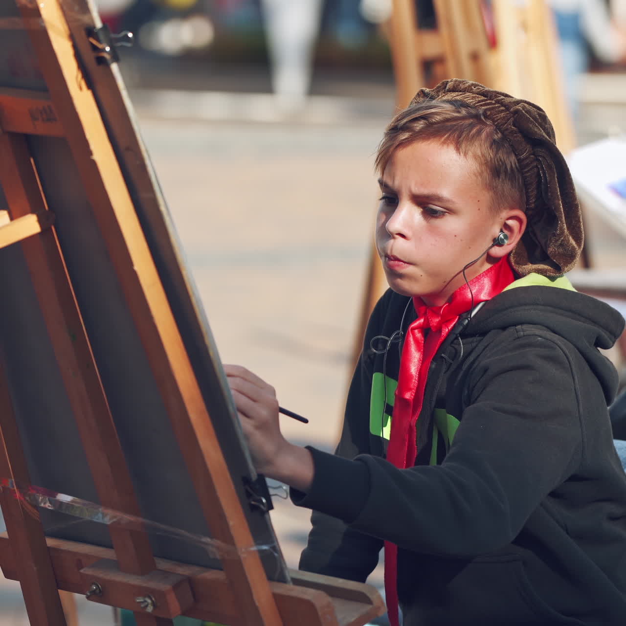 Serious boy is painting on easel in the city. Portrait of a kid artist practices his skills in drawing with brush on the busy city background. City fair.