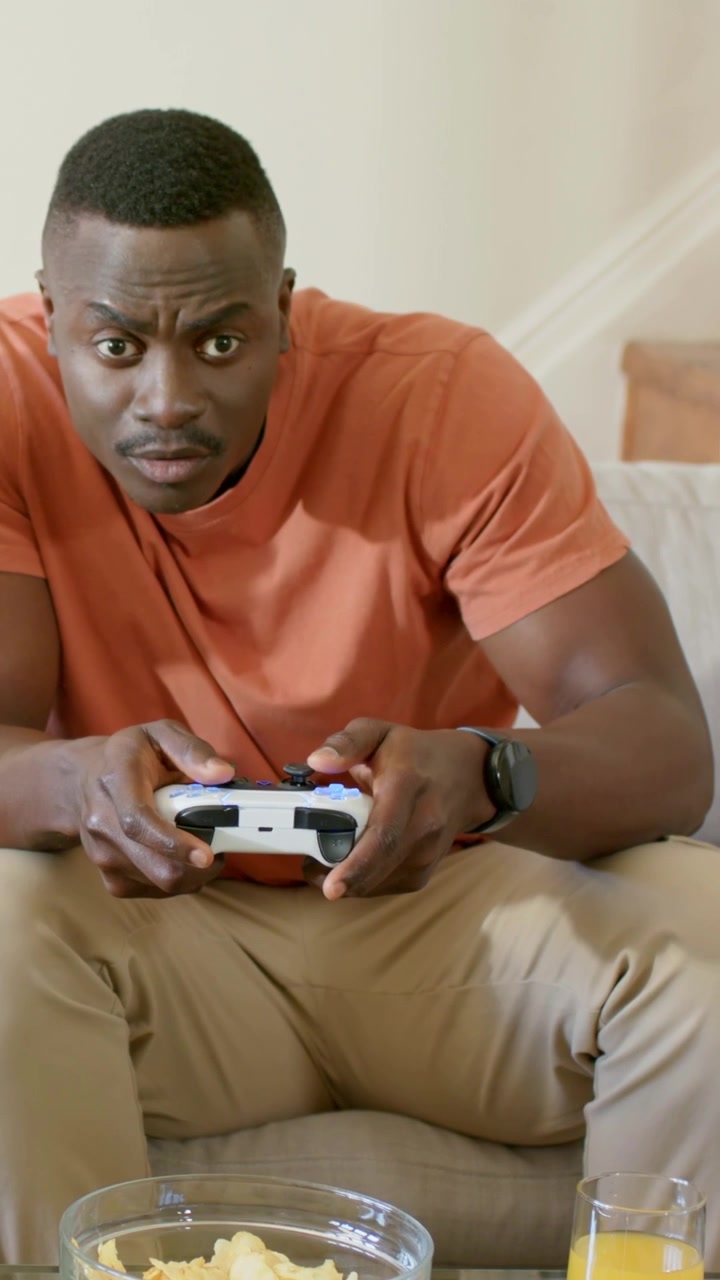 Vertical video: Reacting to game start man in orange tee gripping white gamepad on couch, chips OJ