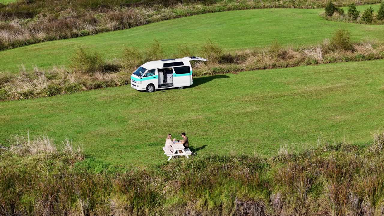 Un drone 4K riprende una coppia seduta su una panchina che beve caffè in un campo verde accanto a un lago in un campeggio libero in Nuova Zelanda. Le acque calme e la vegetazione lussureggiante creano una scena pacifica