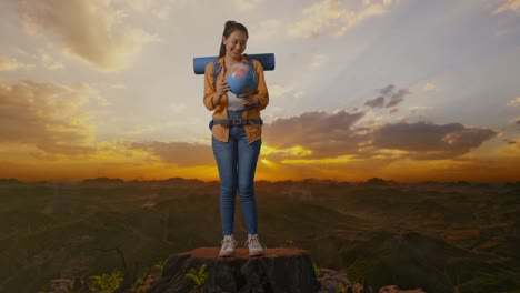 cuerpo lleno de excursionista femenina asiática con mochila de montañismo sosteniendo el globo mundial en sus manos y sonriendo mientras estaba de pie en la cima de la montaña durante la hora de la puesta del sol