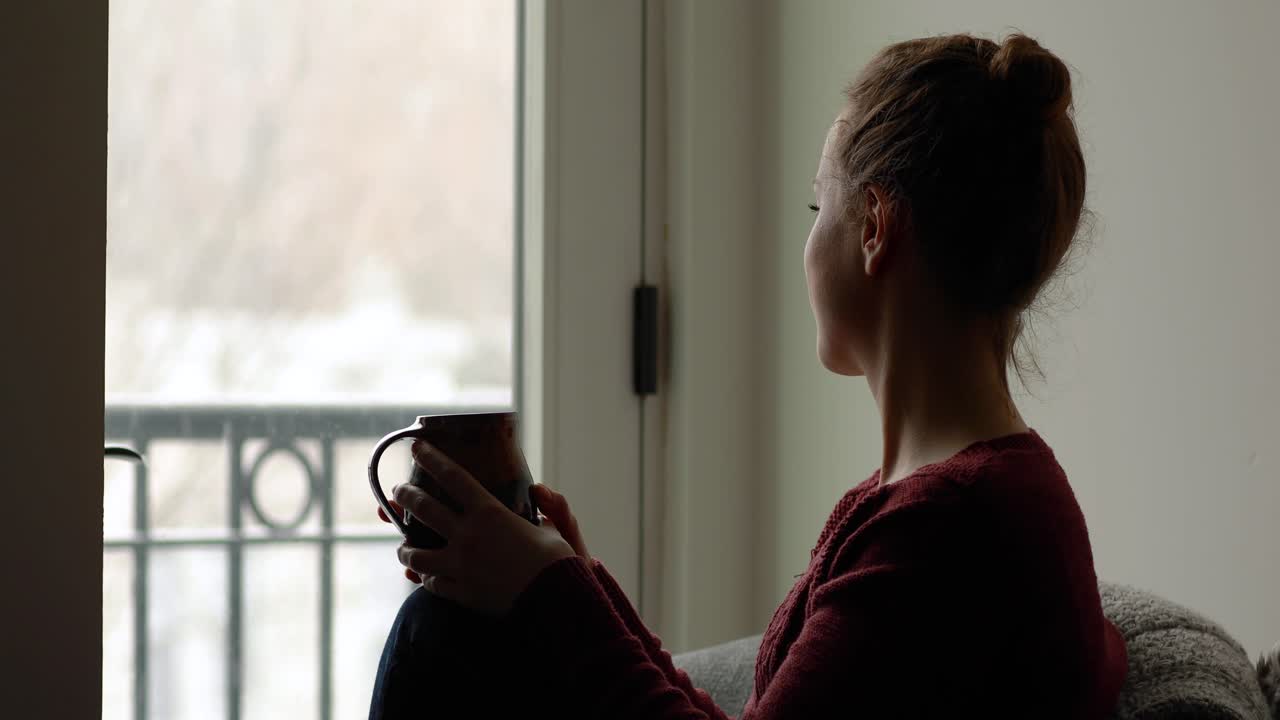Una giovane donna ride, sorseggia una bevanda calda e guarda la fotocamera mentre è all'interno di una casa accogliente, mentre i fiocchi di neve cadono fuori.