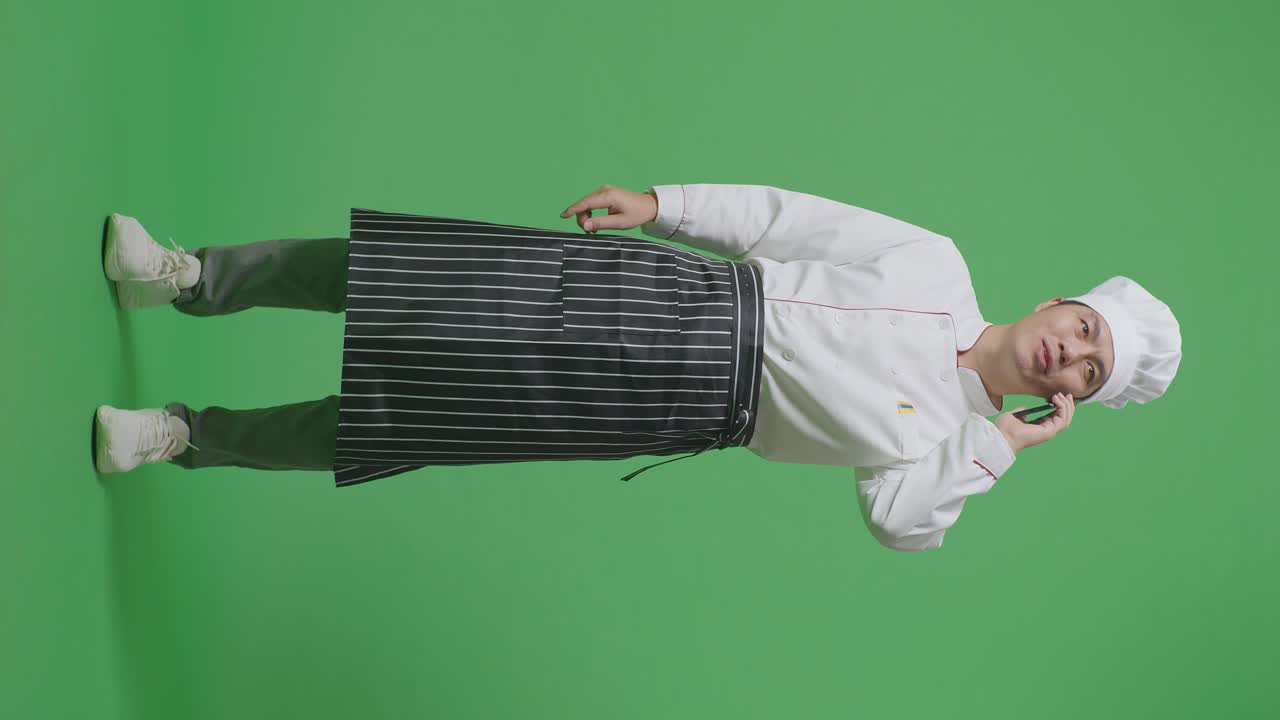 녹색 스크린 배경 스튜디오에 서 있는 동안 스마트폰으로 대화하고 주변을 둘러보는 아시아 남자 셰프의 몸이 가득 차 있습니다.