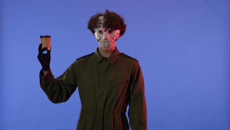 man in white mask with cup of coffee shows thumbs up gesture on blue background with red neon light