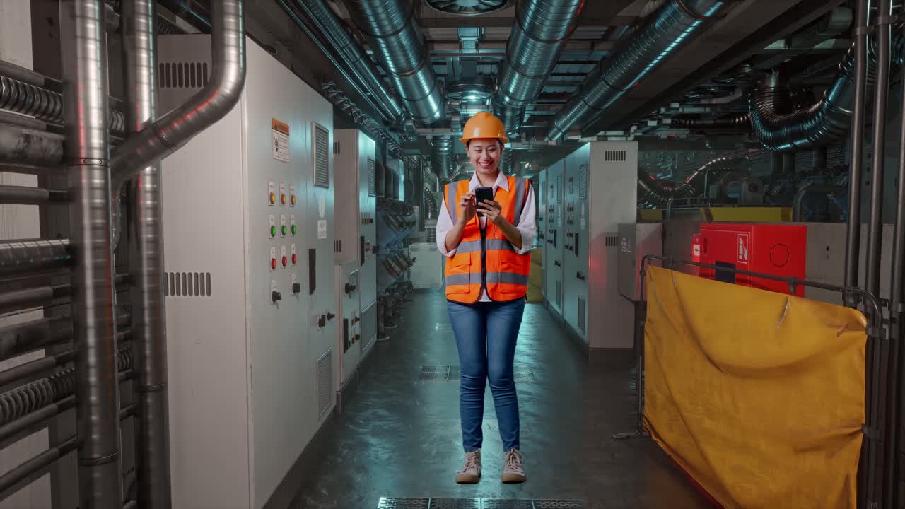 Full Body Of Asian Female Engineer With Safety Helmet Using Smartphone And Looking Around While Standing In Engine Control Room, Work Of Electrical Generators