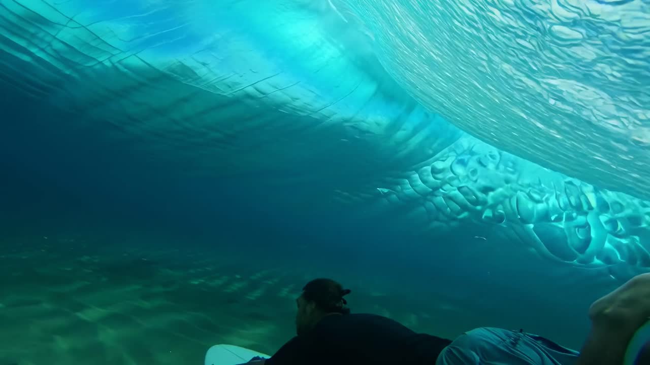 Vídeo subaquático filmado de baixo, capturando um surfista deslizando por ondas azuis vibrantes