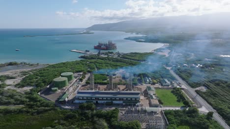 Aerial-backwards-shot-of-smoke-is-rising-at-Electrical-substation-in-Azua-in-front-of-Caribbean-Sea-,-Dominican-Republic