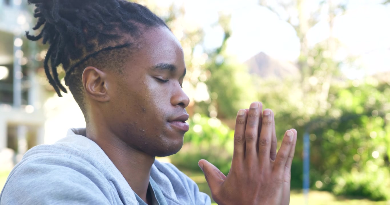 Homme africain adulte levant les bras, paumes vers le bas, et priant dans un parc ensoleillé, avec un sweat à capuche gris et une montre connectée