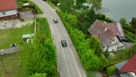 an aerial view of a road alongside a lake, with houses and green foliage lining the sides with cars on the road