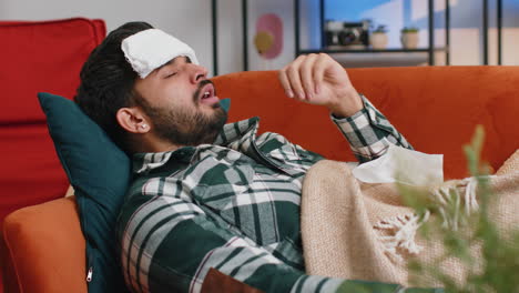 Sick-ill-indian-man-suffering-from-cold-or-allergy-lying-on-home-sofa-sneezes-wipes-snot-into-napkin