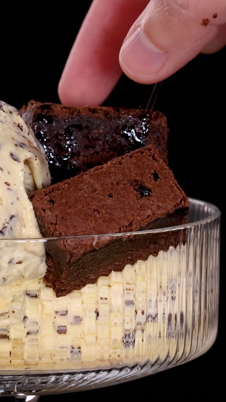 Mano che posiziona un brownie al cioccolato su gelato alla vaniglia con gocce di cioccolato in una ciotola di vetro, sfondo nero