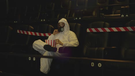 portrait of a man in white protection costume and respirator sitting alone at the cinema taking popcorn